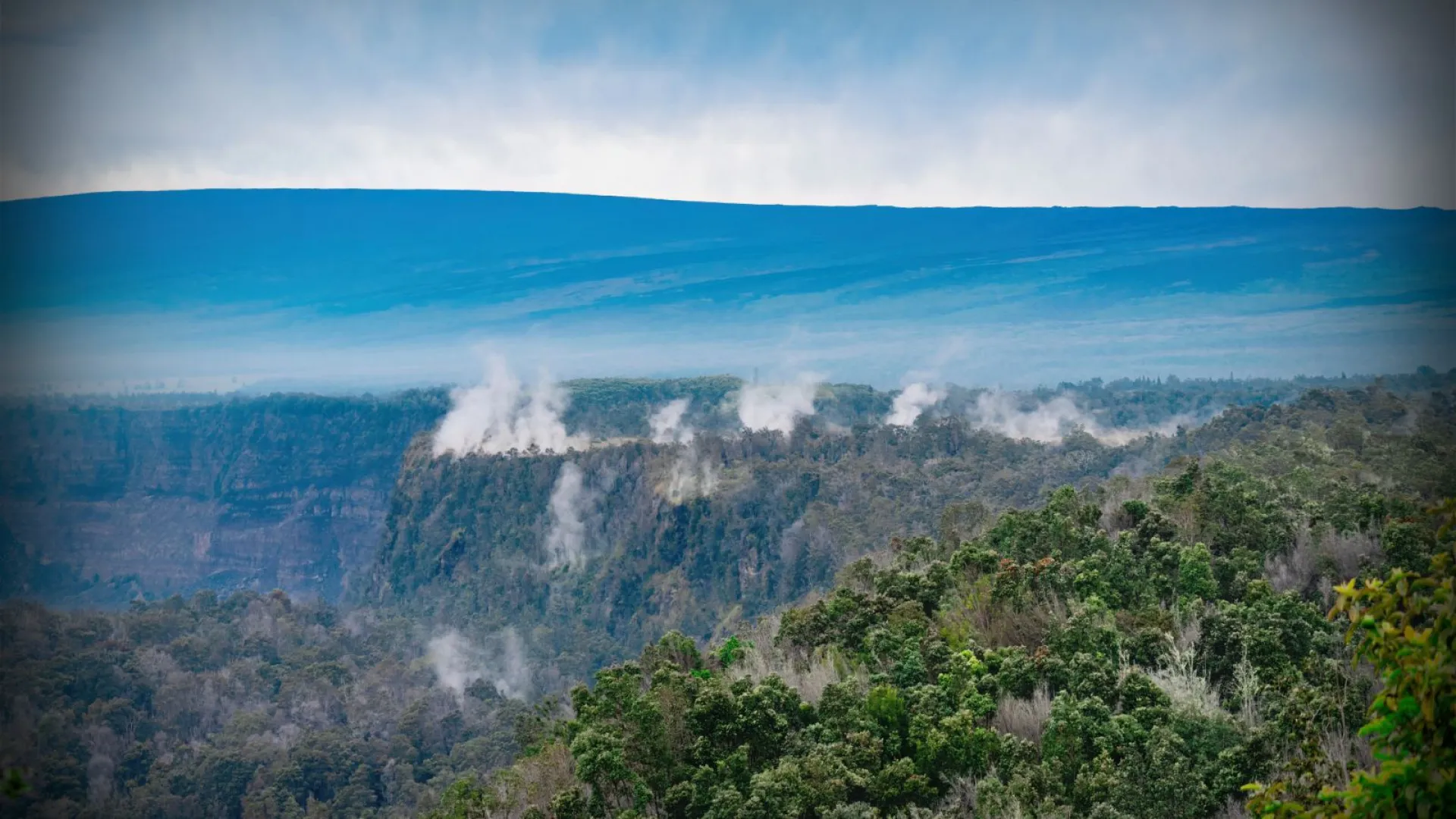 Steam rising from a lush green forest with distant mountains.